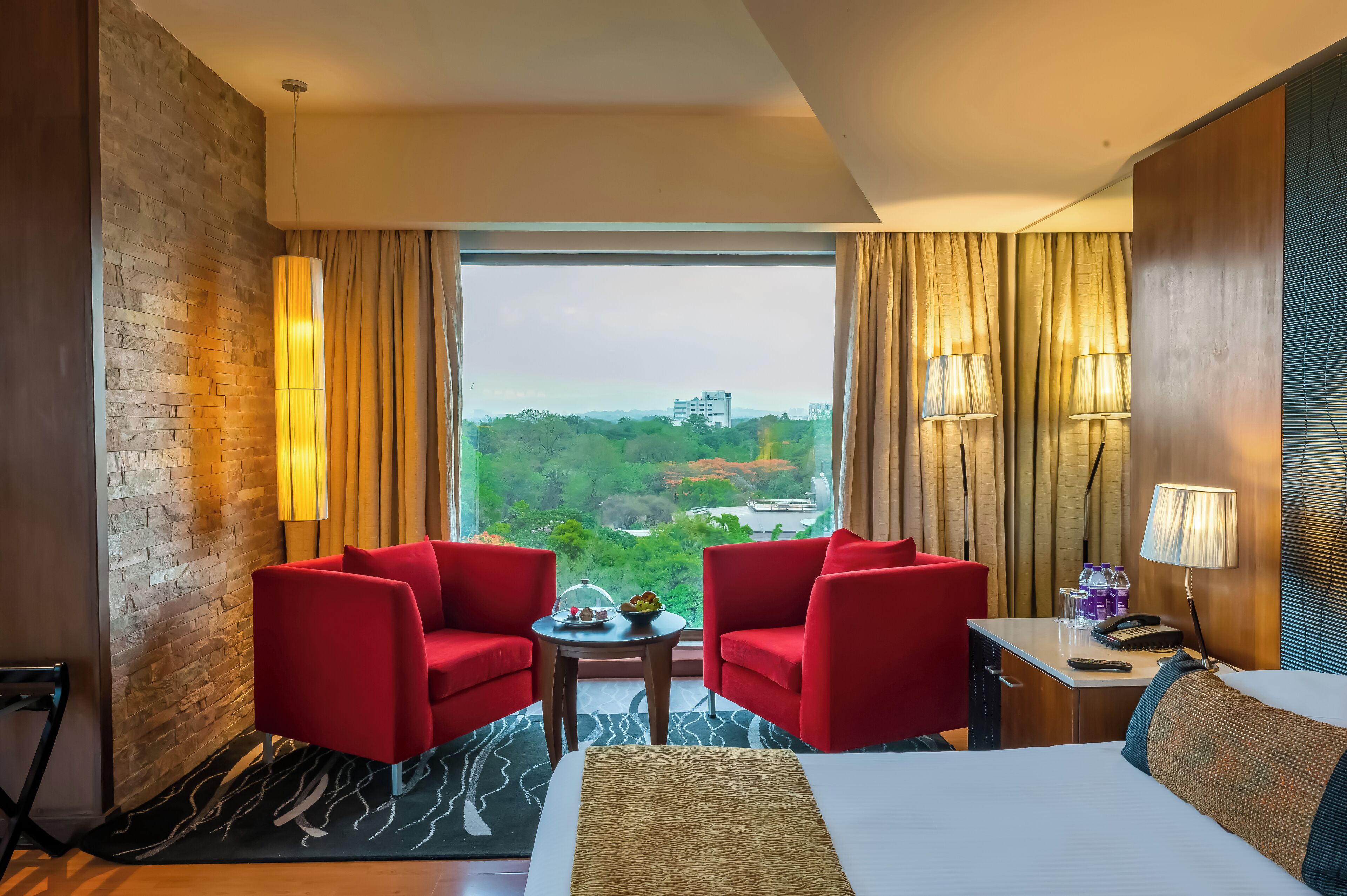 Habitación de hotel con una cama grande, dos sillones rojos, una mesita con lámpara y vistas a zonas verdes y edificios.