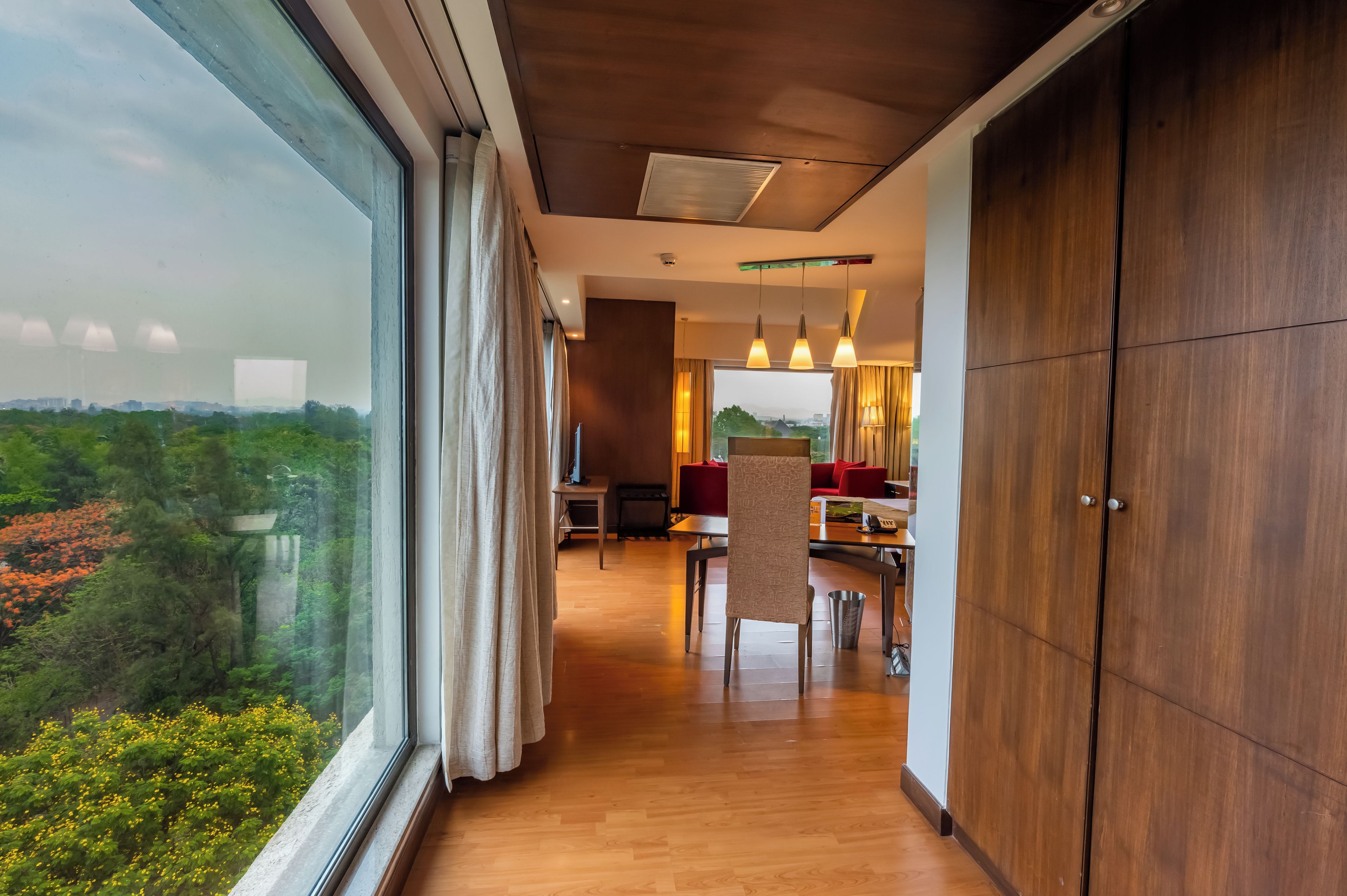 A modern interior with a wooden floor, a dining area with red chairs, and a view of greenery outside.