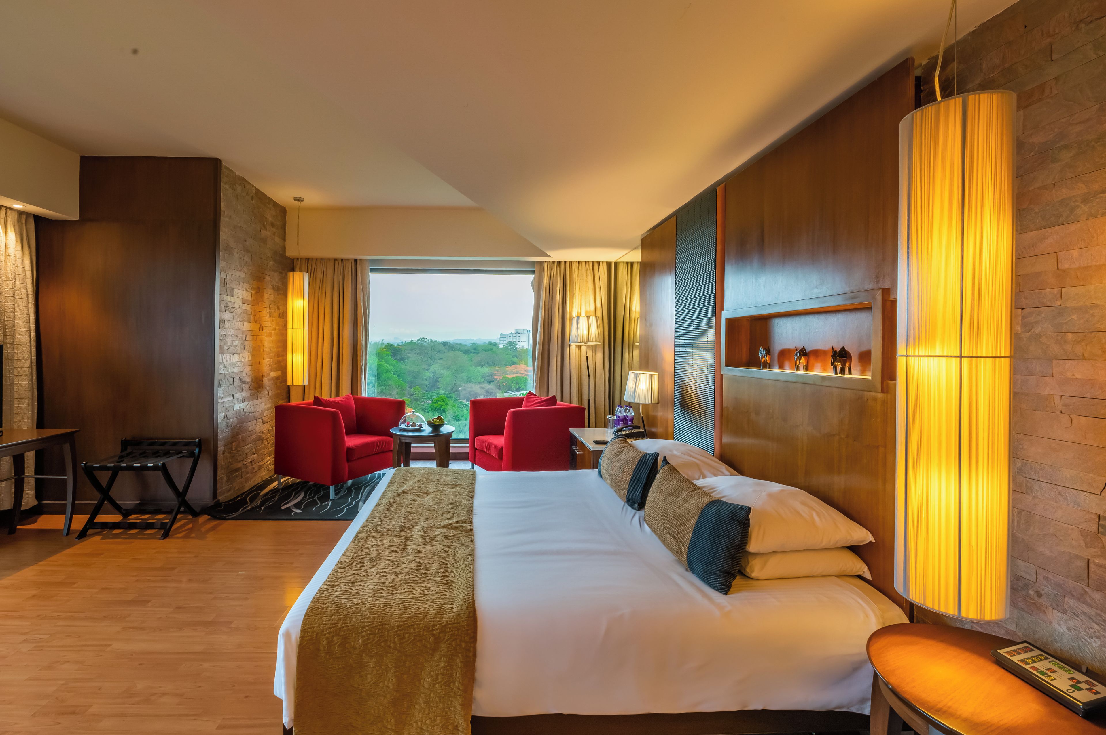 A hotel room with a large bed, two red armchairs, a small table, and a view of the outdoors.