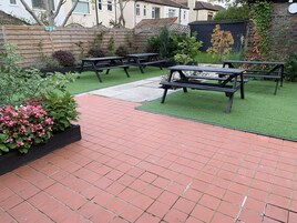A patio area with picnic tables and artificial grass, surrounded by a wooden fence and various plants.