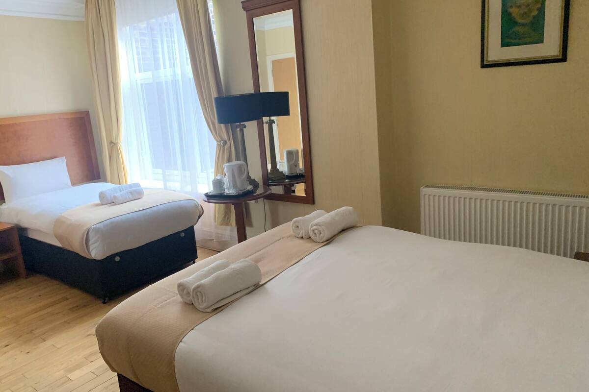 A hotel room with two beds, a bedside table with a lamp, a radiator, and a framed picture on the wall.