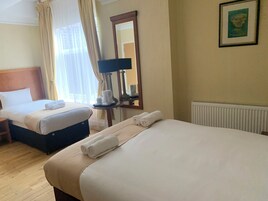 Habitación de hotel con dos camas, una mesita de noche con lámpara, un radiador y un cuadro en la pared.