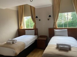 A hotel room with two beds, beige curtains, a nightstand, and a view of greenery outside.