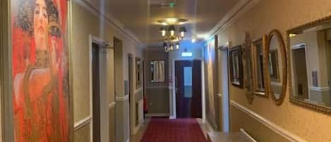 A hallway with a red carpet, a bench, a painting on the left, and a chandelier.