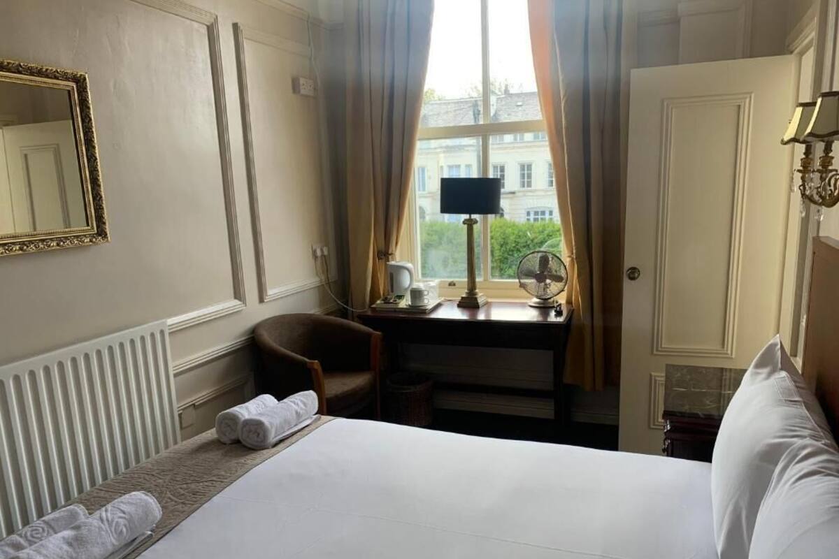A hotel room with a bed, a desk with a lamp, a chair, a window with curtains, and a radiator.
