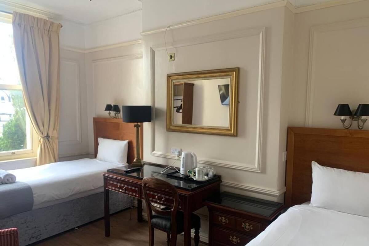 A hotel room with two single beds, a wooden desk with a chair, a small table with a lamp, and a mirror on the wall.