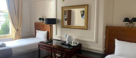 Une chambre d’hôtel avec deux lits simples, un bureau en bois avec une chaise, une petite table avec une lampe et un miroir au mur.