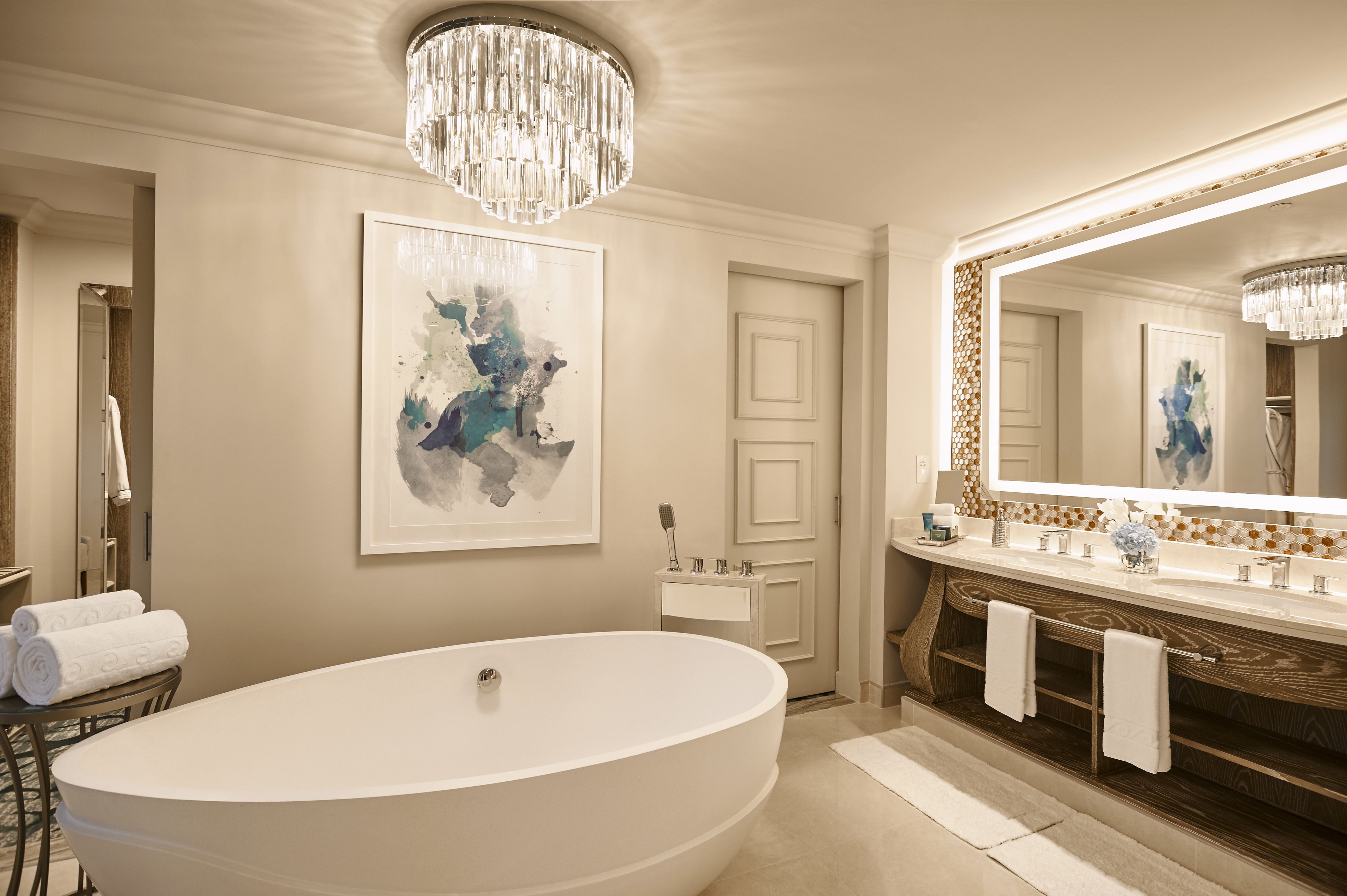 A modern bathroom with a large freestanding bathtub, a large mirror with a decorative frame, and a chandelier.