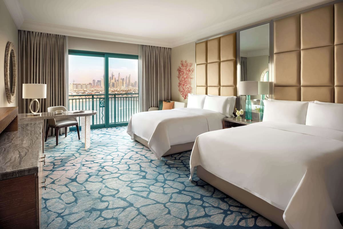 A hotel room with two beds, a desk, a chair, a large window with a city view, and a patterned carpet.