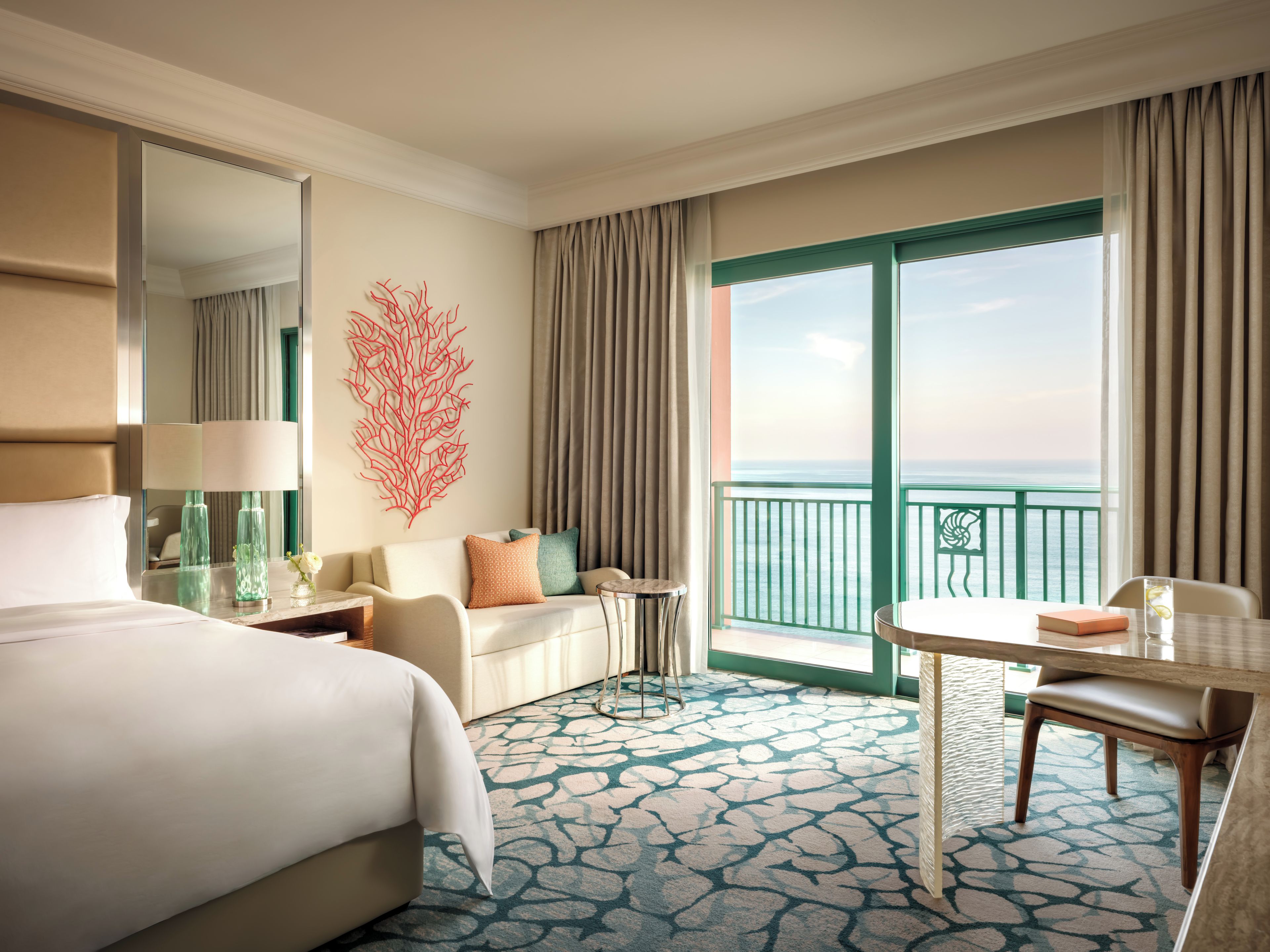 A hotel room with a bed, a seating area, a balcony with a view, and a decorative wall piece.
