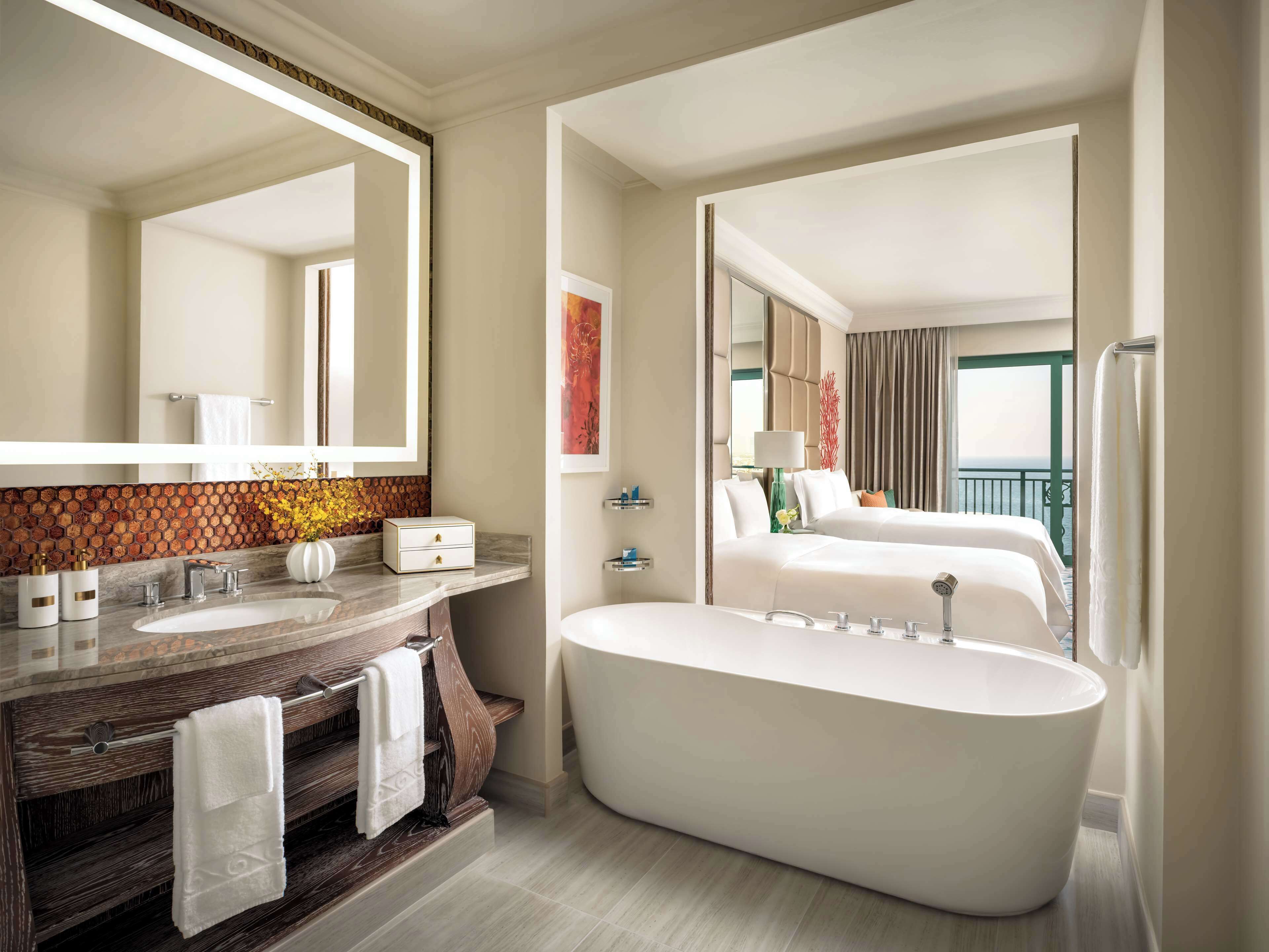 A modern bathroom with a large bathtub, a vanity with a mirror, and a separate bedroom with a bed and a view of the balcony.