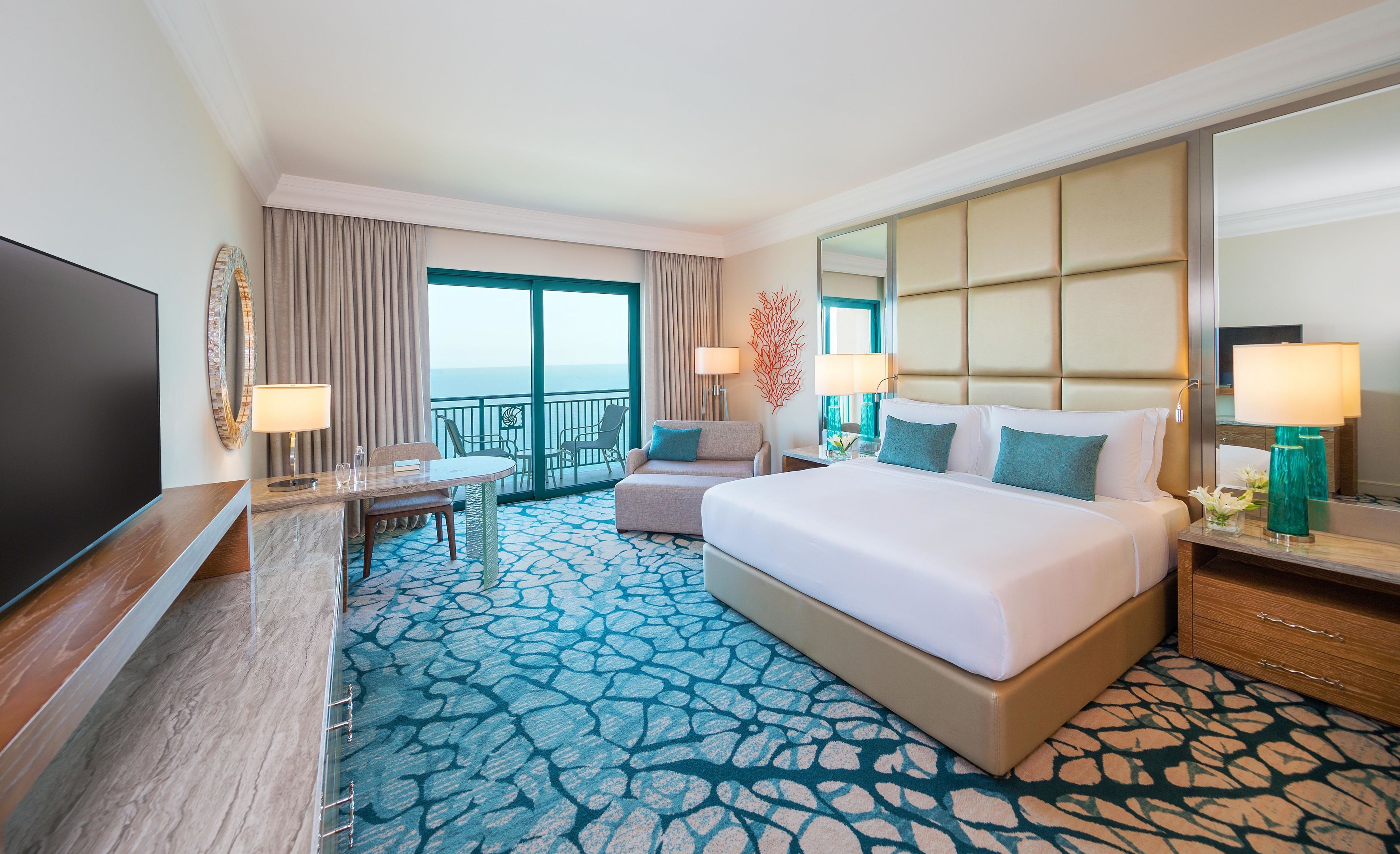 A hotel room with a large bed, a desk with a TV, a balcony with a view, and a patterned carpet.