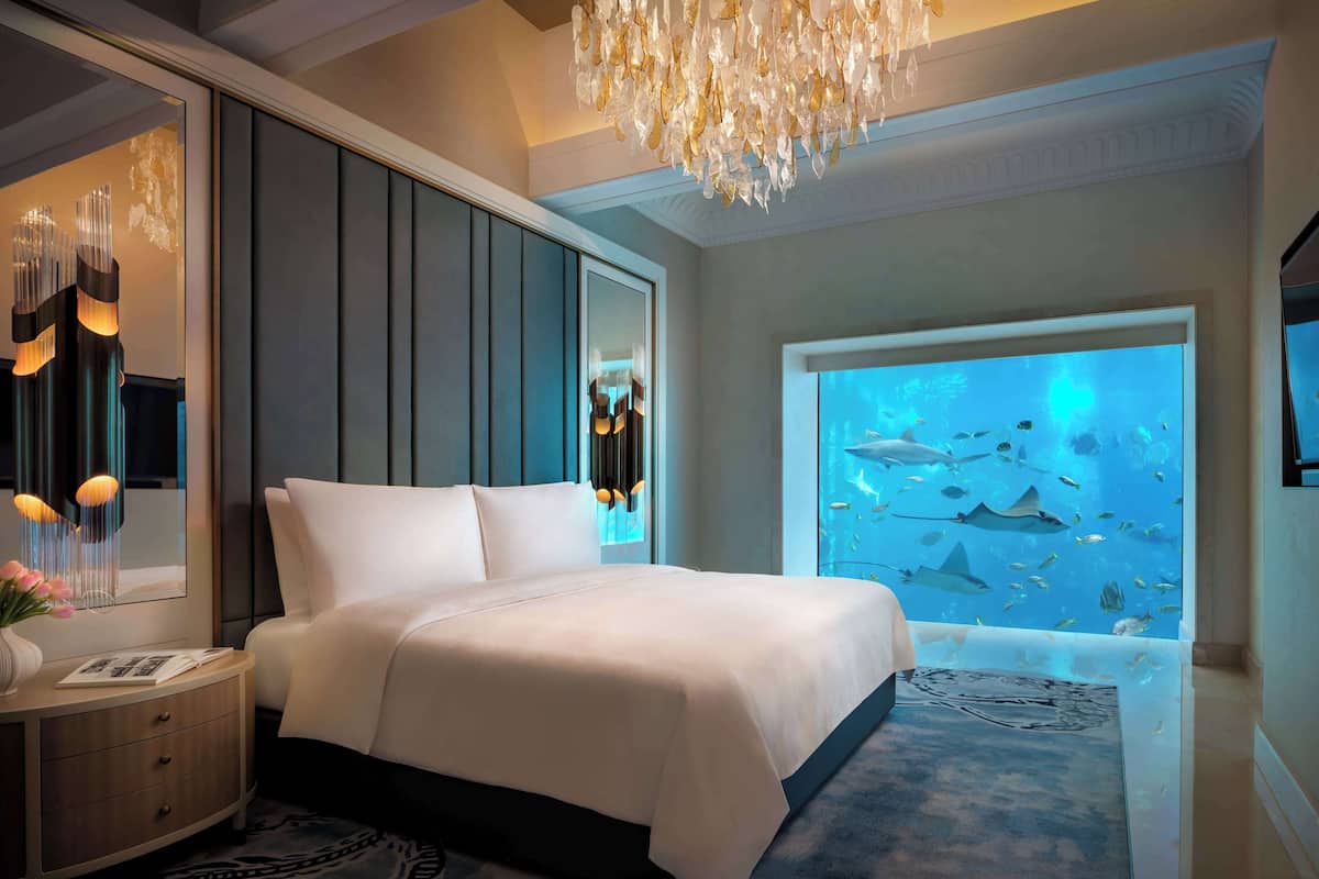 A bedroom with a large bed, a nightstand, a mirror, a chandelier, and an aquarium wall.