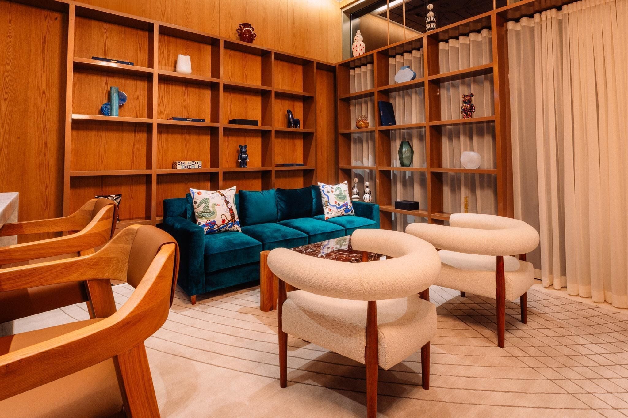 A cozy living room with a teal sofa, wooden chairs, a bookshelf, and a window with curtains.