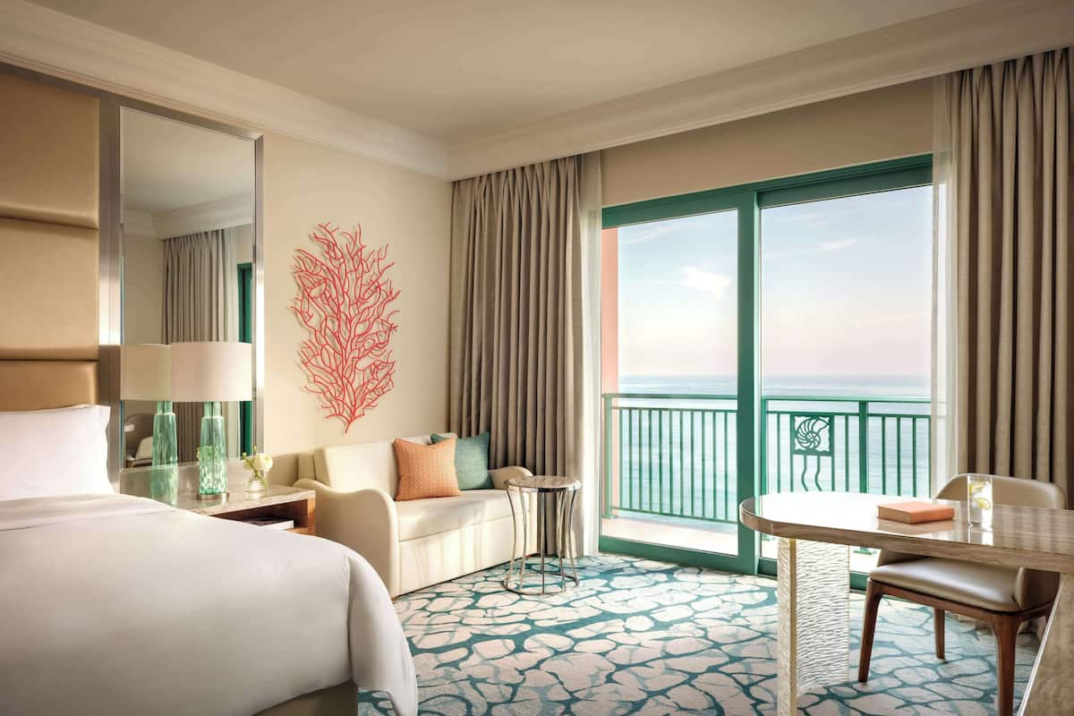 A hotel room with a bed, a seating area with a table and chairs, a balcony with a view, and a decorative wall piece.