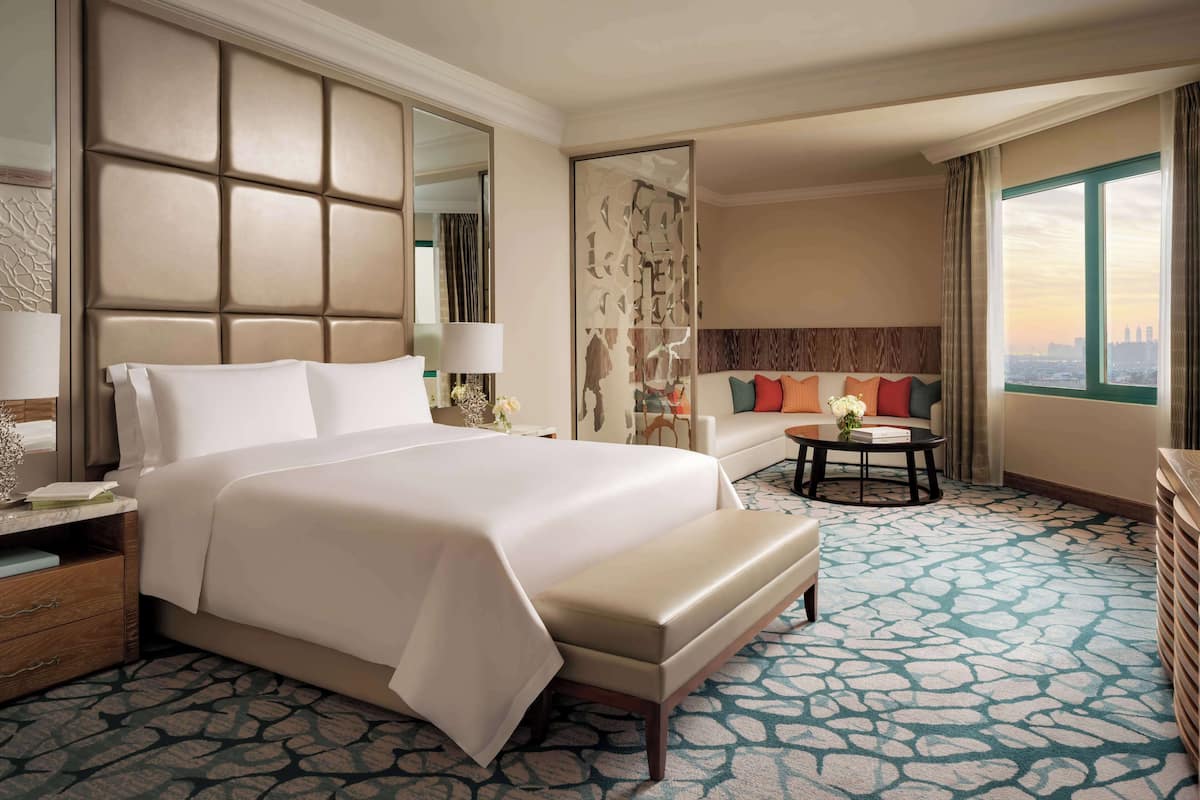 A hotel room with a large bed, a sofa, a small table, a mirror, and a window with a city view.