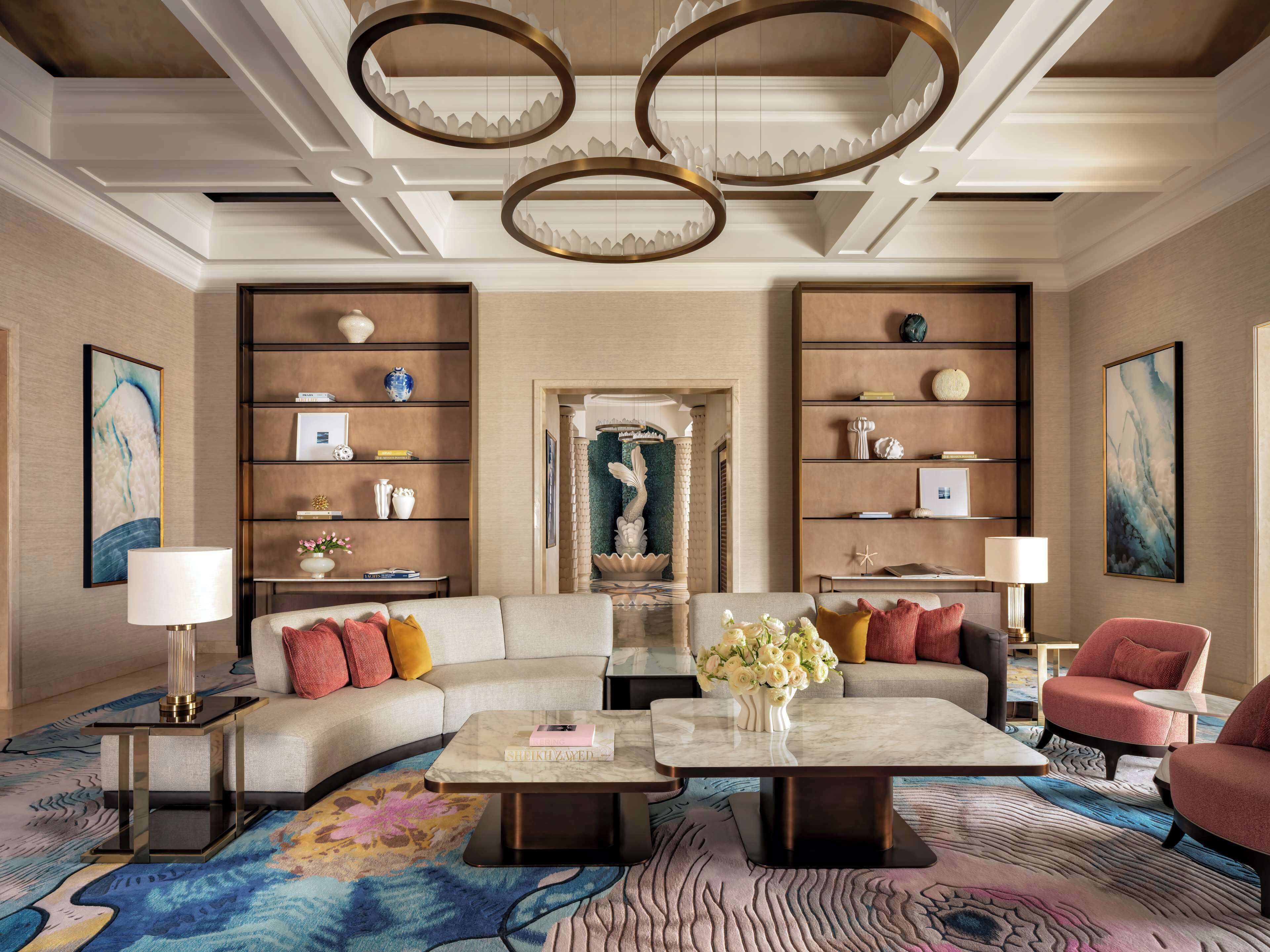 A modern living room with a sofa, coffee table, and armchairs, featuring a decorative mirror, bookshelves, and a floral centerpiece.