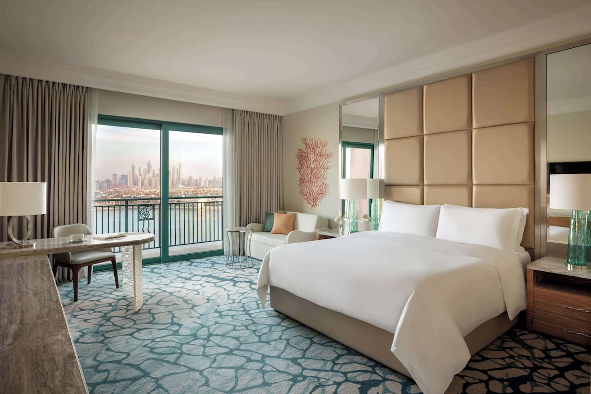 A hotel room with a large bed, a desk, a chair, a sofa, a large mirror, and a view of a cityscape.