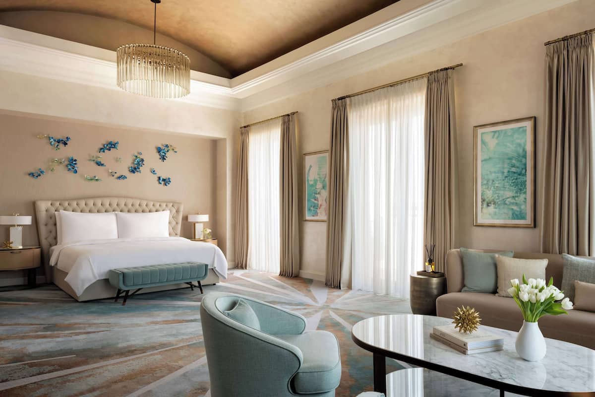 A spacious hotel room with a large bed, a sitting area with a sofa and armchairs, a round coffee table, and a chandelier.