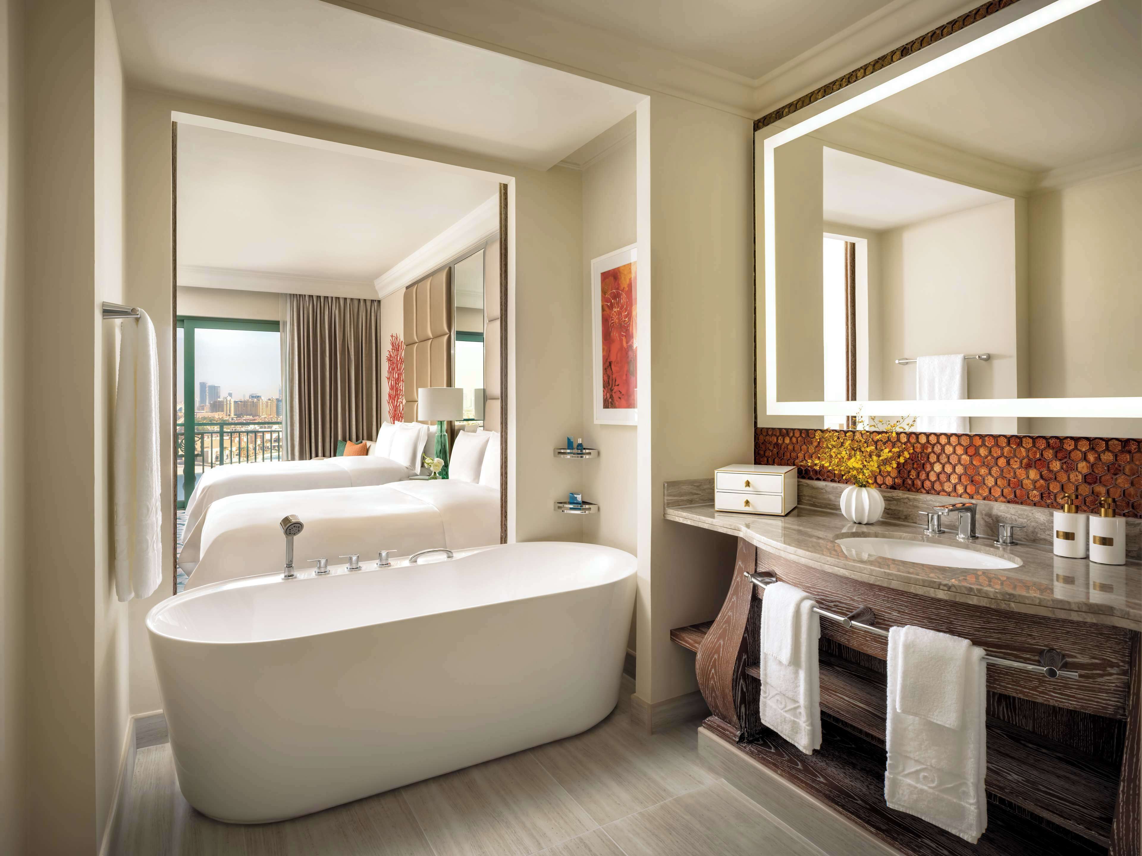 A modern hotel bathroom with a large bathtub, two beds, a vanity with a mirror, and a city view.