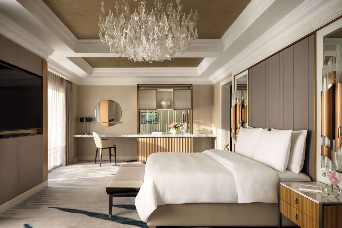 A modern hotel room with a large bed, a desk, a chair, a television, and a chandelier.