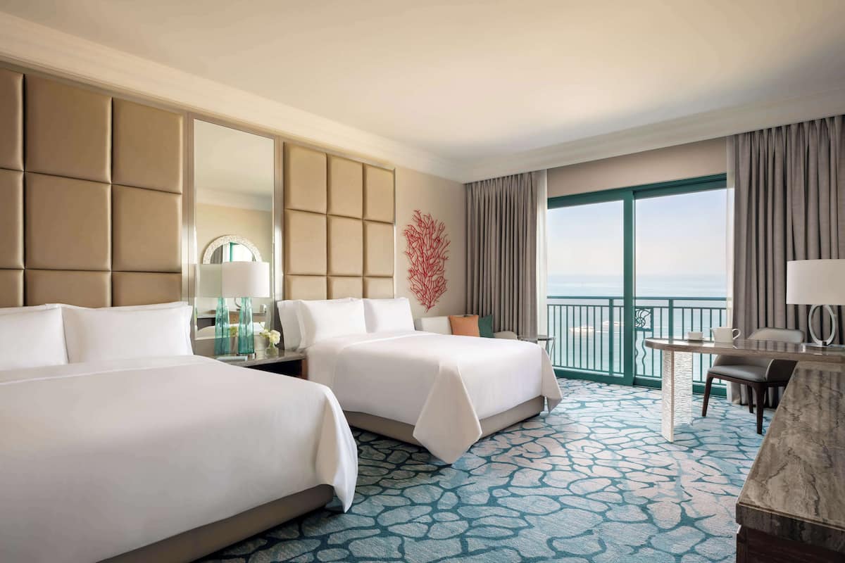 A hotel room with two beds, a desk, a chair, a large window with a view, and a decorative wall piece.