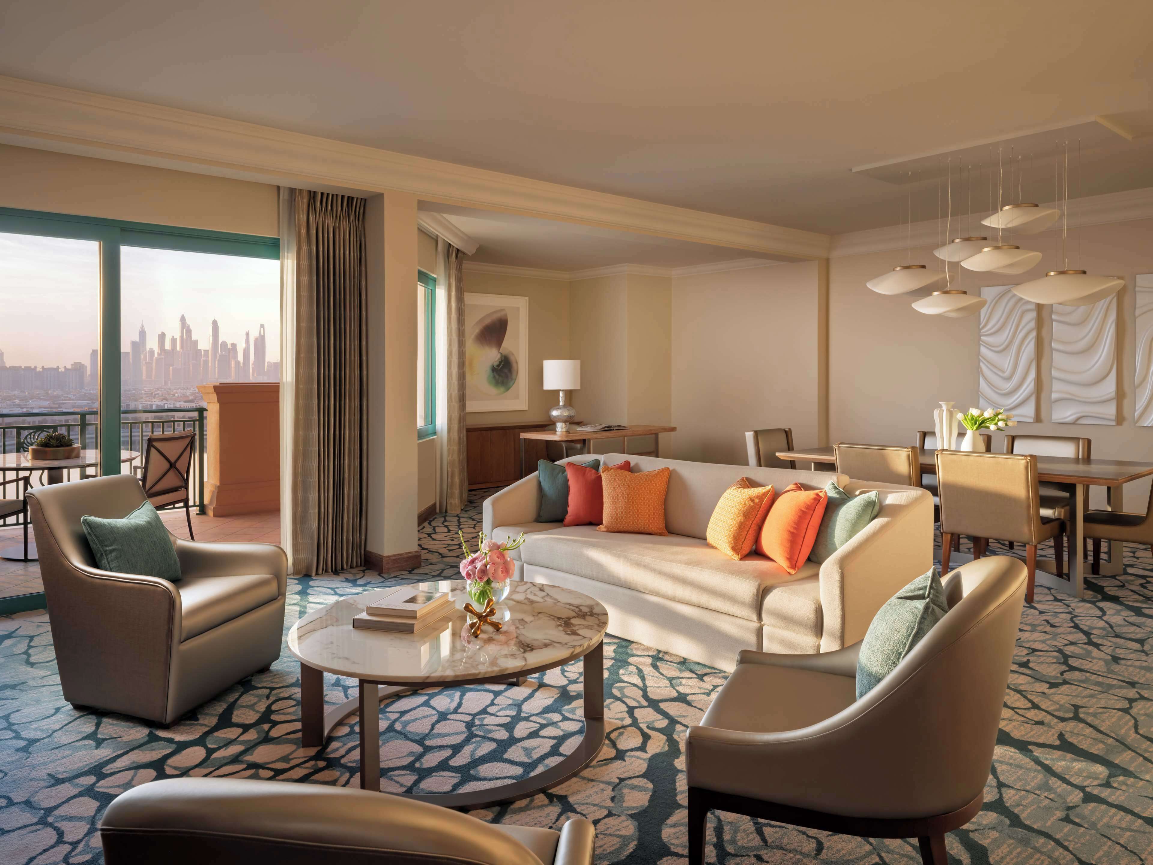 A hotel room with a sofa, chairs, a dining table, a coffee table, and a view of a city skyline.