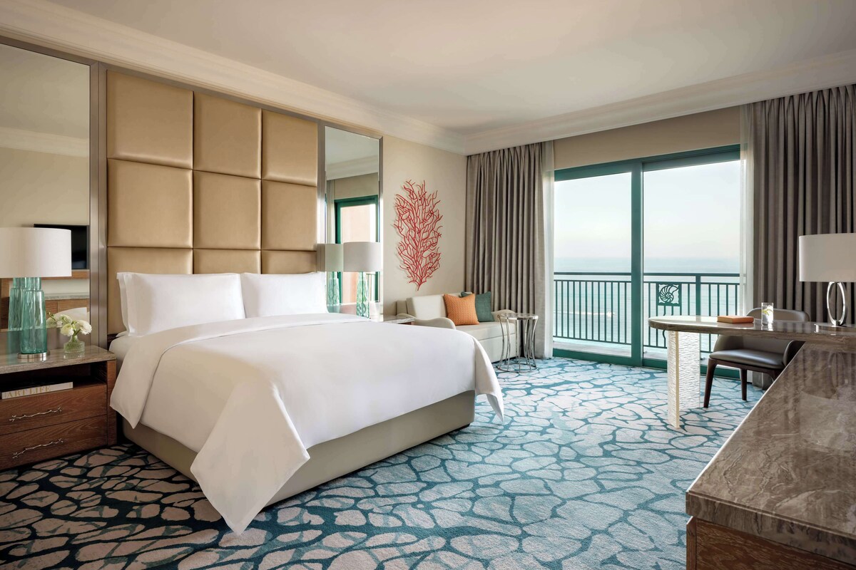 A hotel room with a large bed, a desk, a chair, a balcony with a view, and a decorative wall piece.