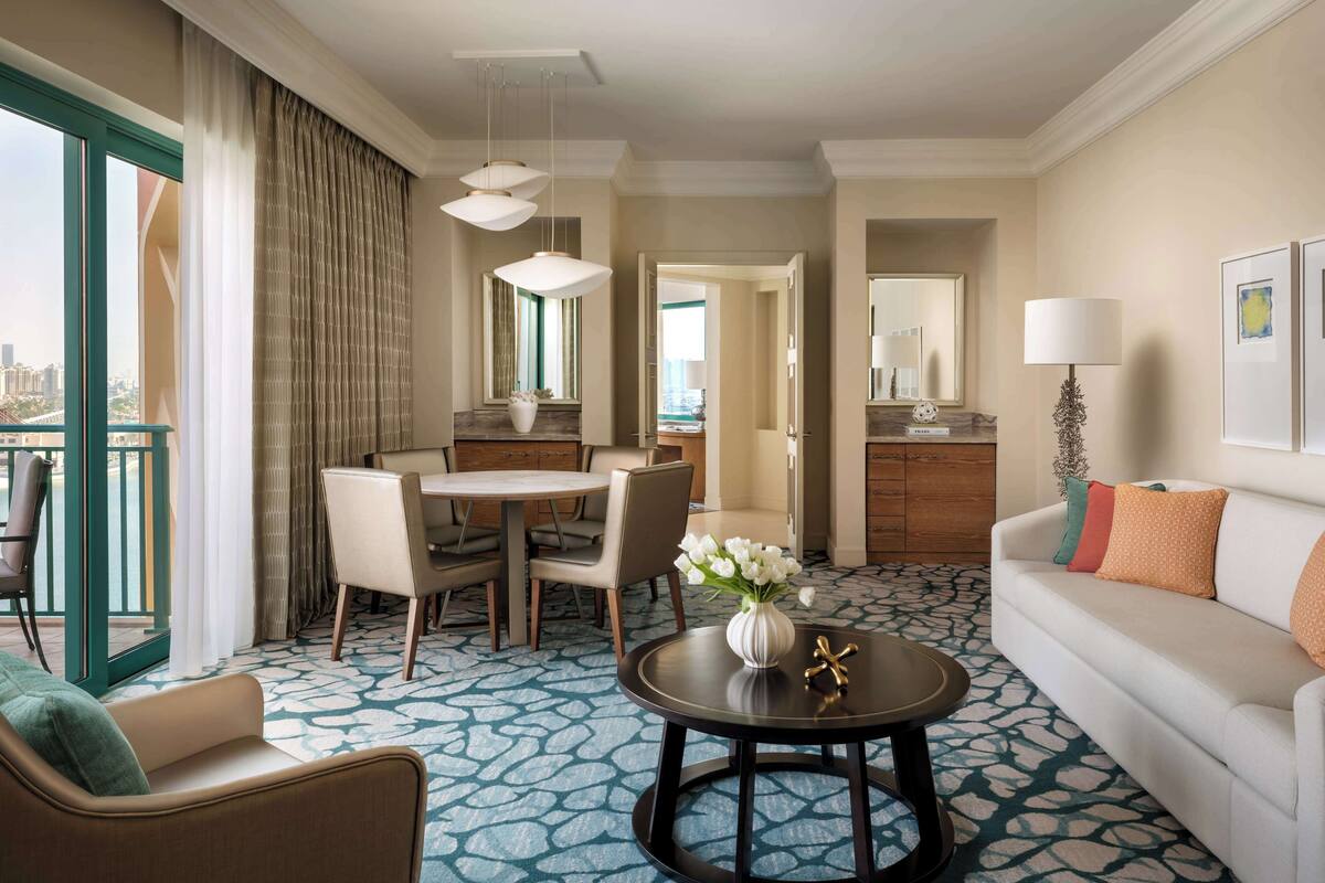 A hotel room with a dining area, a sofa, a coffee table with flowers, and a view of the city.