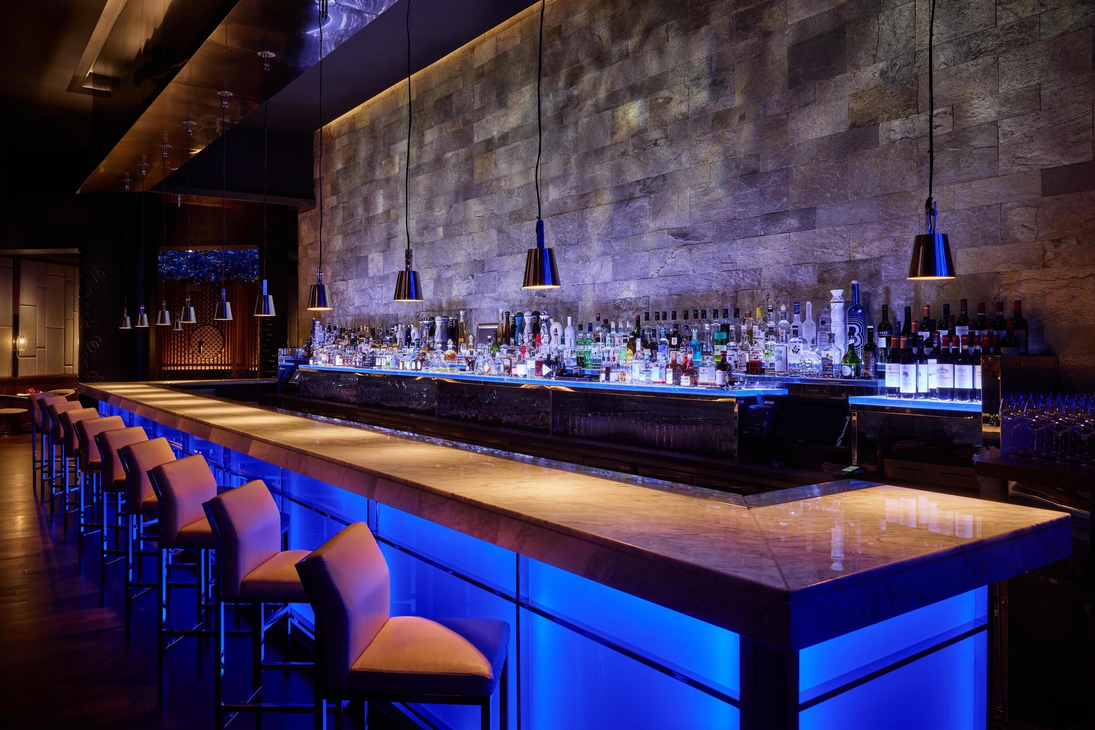 A bar area with a long counter, multiple stools, and a variety of bottles.