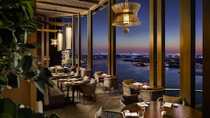 A restaurant with tables and chairs, large windows with a view of a cityscape and water, and hanging lamps. - Atlantis, The Palm (Dubai)