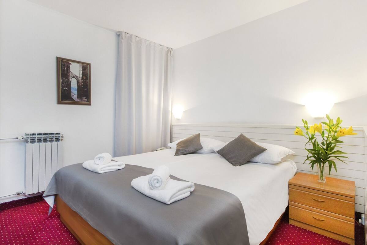 A hotel room with a bed, two pillows, a bedside table with a vase of flowers, a radiator, and a painting on the wall.