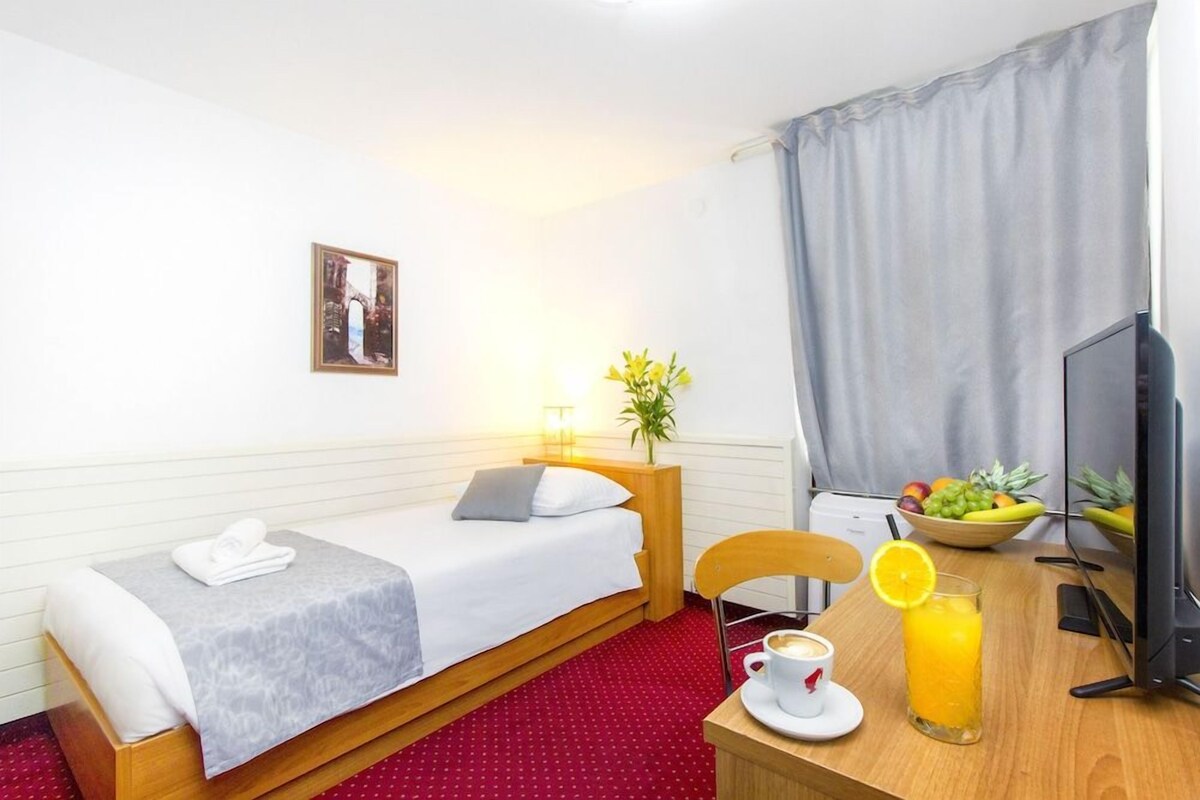 A hotel room with a bed, a desk with a chair, a television, a coffee cup, a glass of orange juice, and a fruit bowl.
