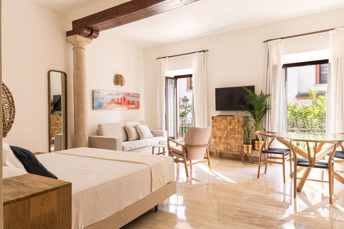 Amplia habitación de hotel con una cama grande, un sofá, una mesa de comedor con sillas, televisión y un balcón con vistas.
