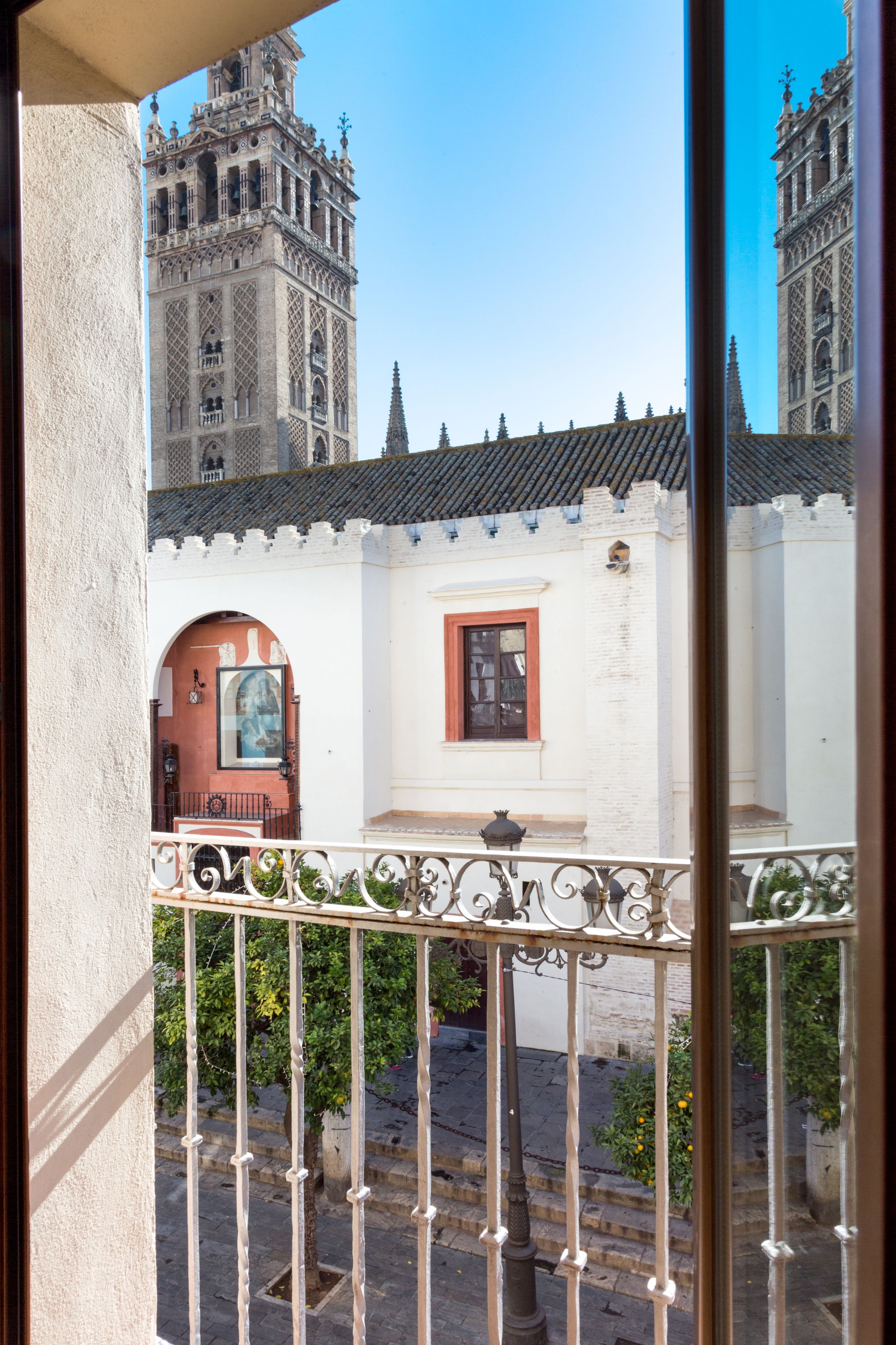 A balcony with ornate metal railings, a view of a building with a red door and a window, and a clear blue sky.