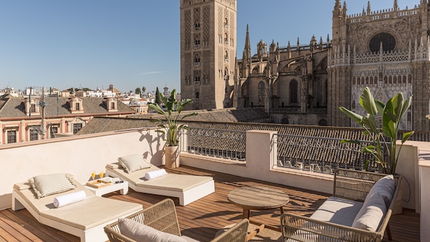 A rooftop terrace with lounge chairs, a wooden deck, and a view of a historic cathedral with a tall tower. - Puerta Catedral Apartments (Seville)