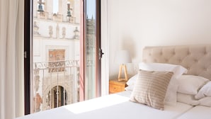 A hotel room with a bed, a nightstand, a lamp, and a view of a building through a glass door. - Puerta Catedral Apartments (Seville)