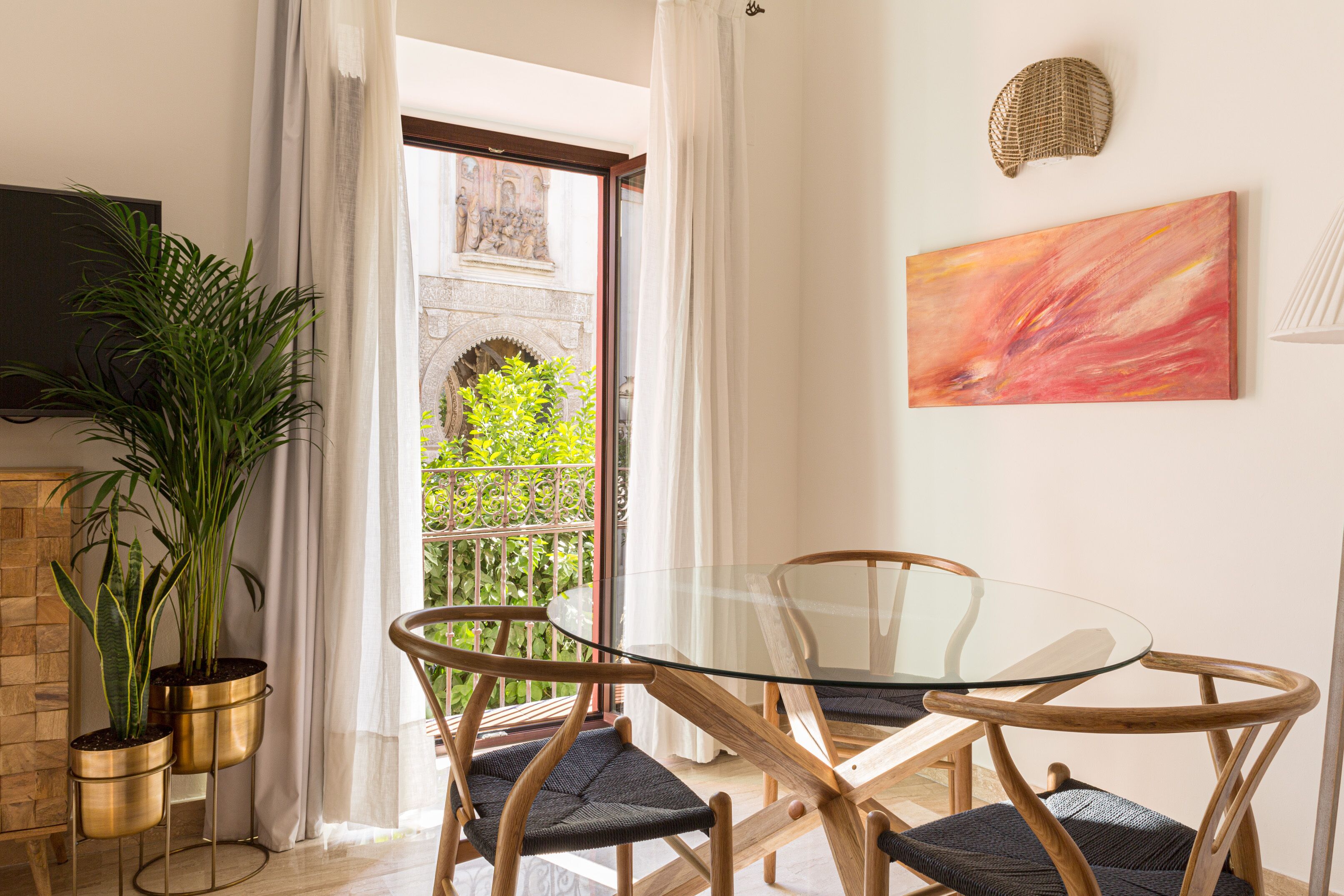 Uma área de refeições moderna com mesa de vidro e cadeiras de madeira, uma planta em vaso e vista para o verde através de uma porta de vidro.