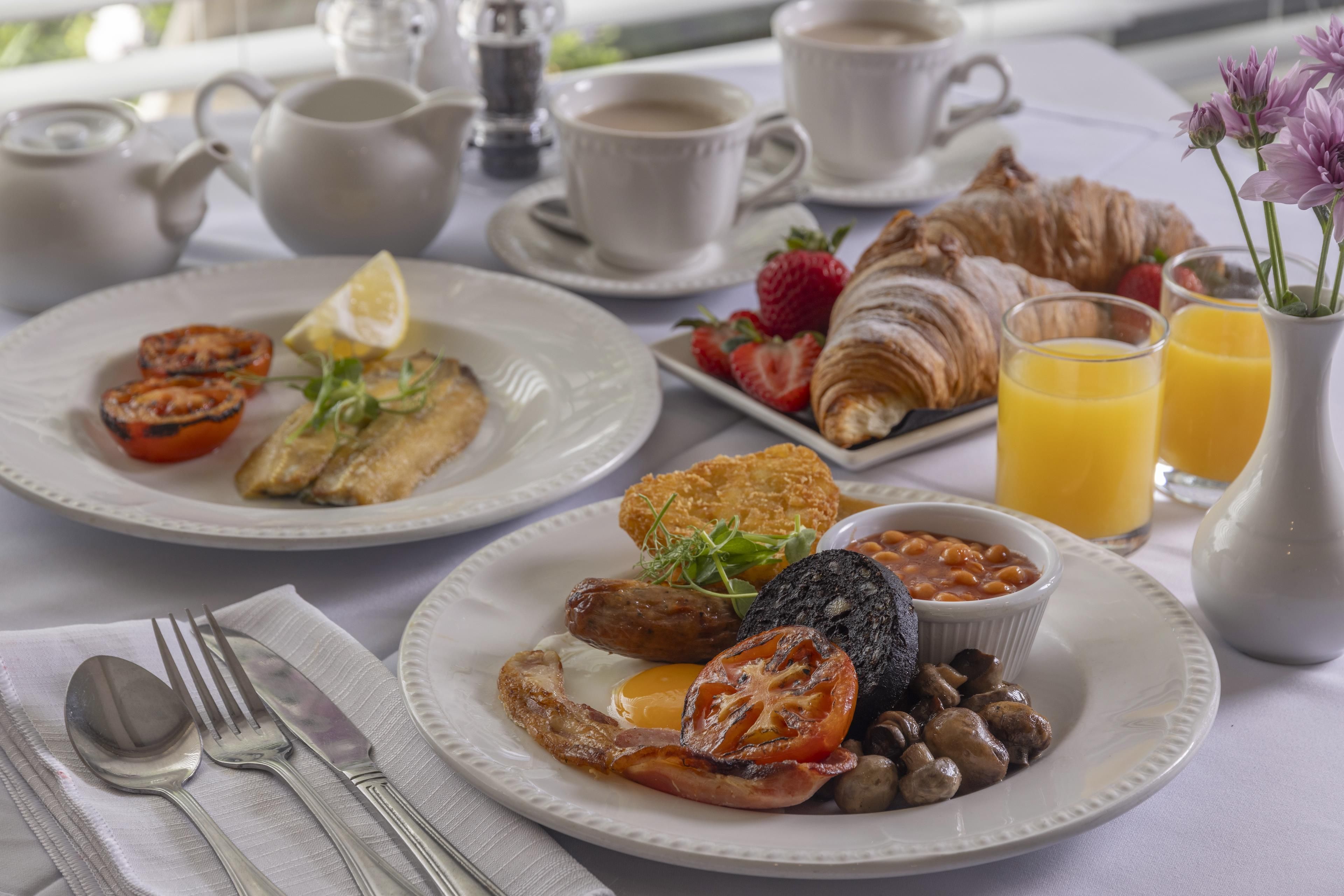 Una mesa de desayuno con huevos, tocino, tomates y champiñones, junto con croissants, fresas y jugo.