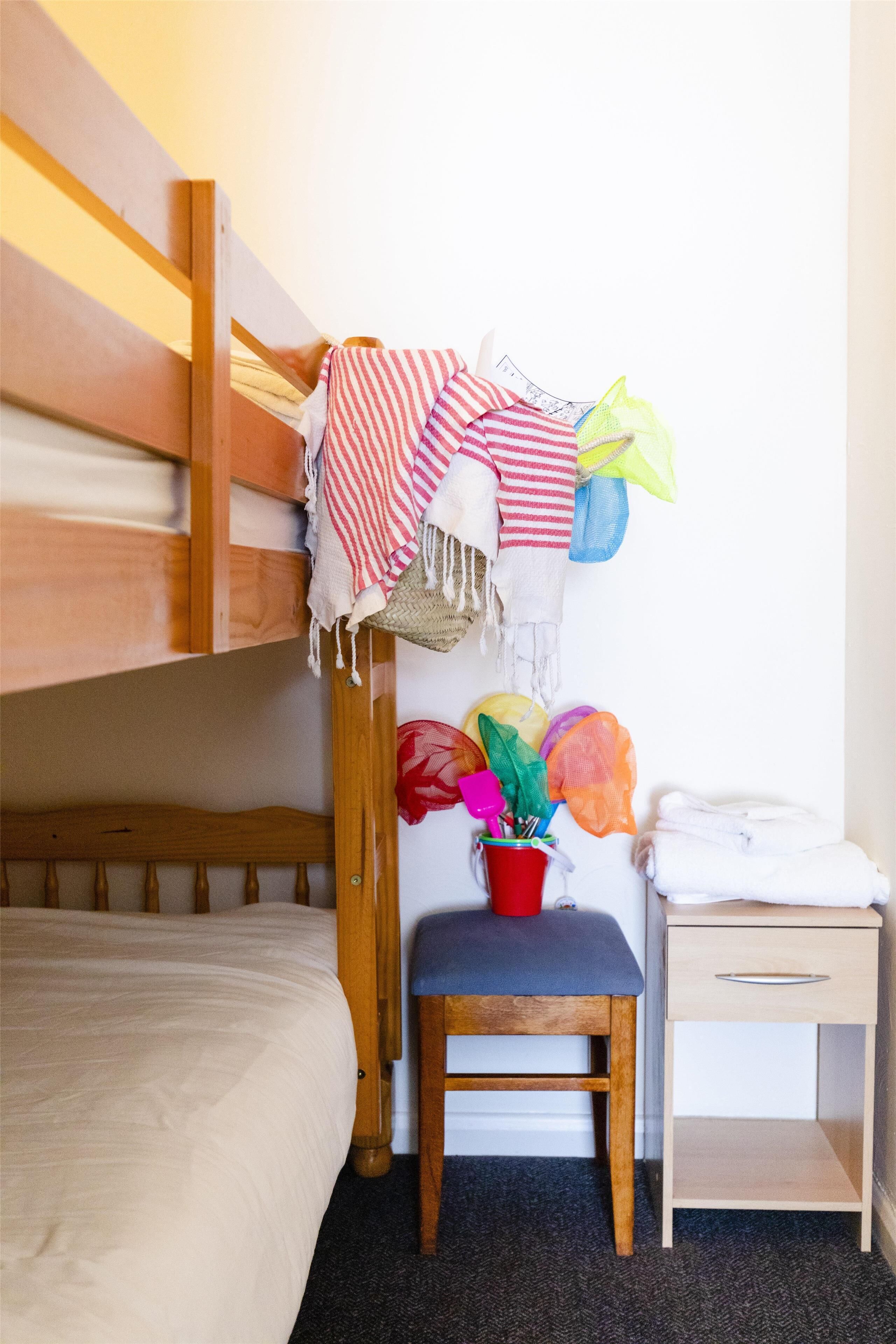 A bunk bed with a wooden headboard, a small wooden table with a red bucket, and a bedside drawer.