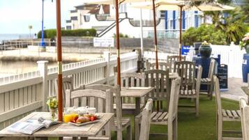 An outdoor dining area with wooden tables and chairs, a white fence, and a view of the water.