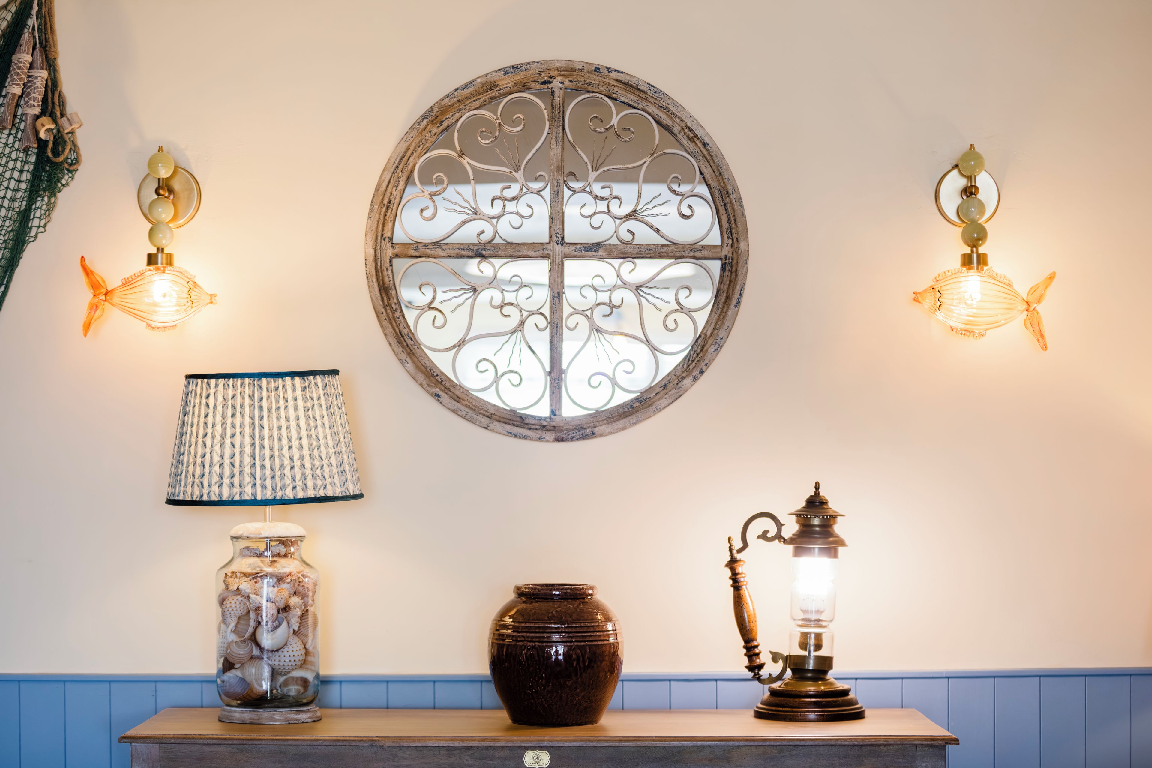 A wall with a decorative metal window, two wall-mounted lights, a lamp, a jar of seashells, and a vintage lantern.