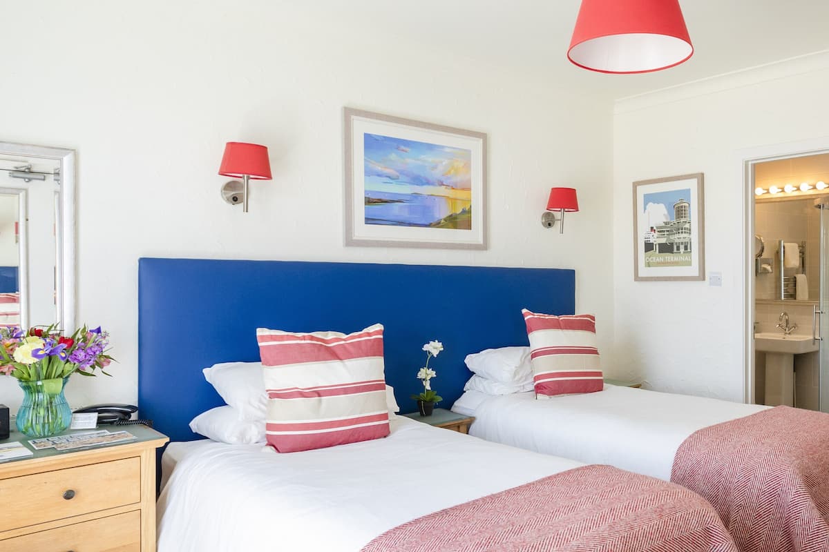 A hotel room with two beds, a wooden wardrobe, a bathroom with a sink, and a painting on the wall.