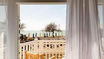 Une vue depuis une fenêtre ornée d’un rideau blanc, donnant sur une plage avec une clôture blanche et un arbre.