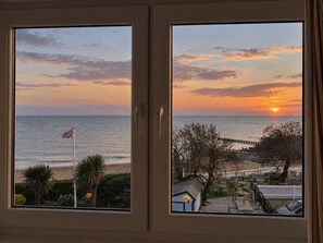 A view through a window with a sunset over the sea, a flag, and a deck area with outdoor seating.