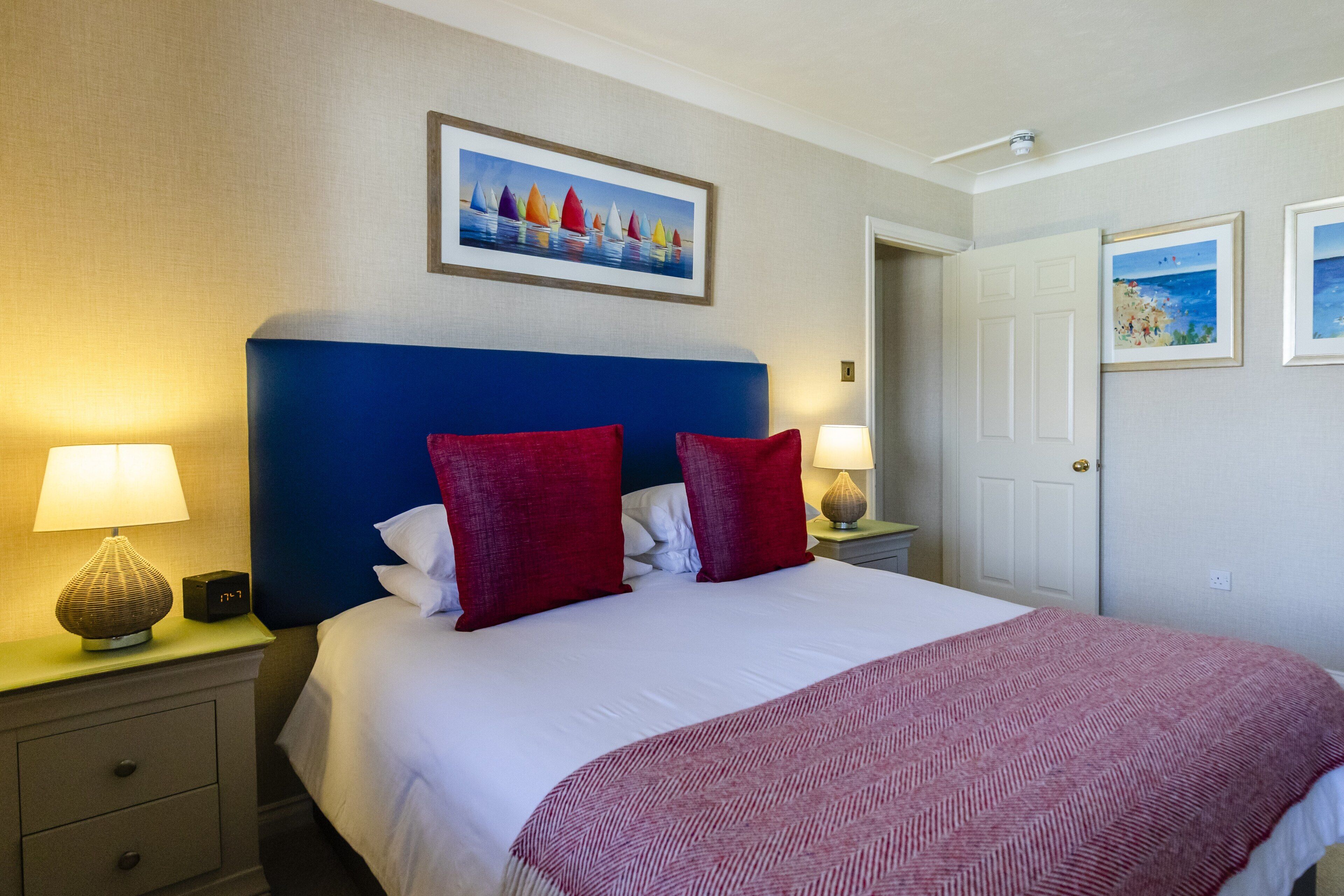 A hotel room with a large bed, red pillows, a white comforter, and a painting above the bed. There is a nightstand with a lamp and a clock.