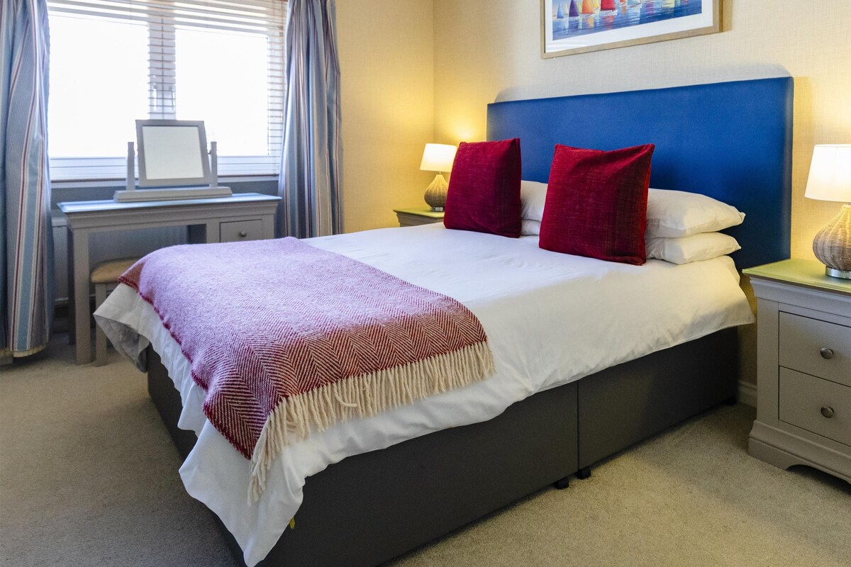 A hotel room with a bed, two bedside tables with lamps, a desk with a computer, and a window with curtains.