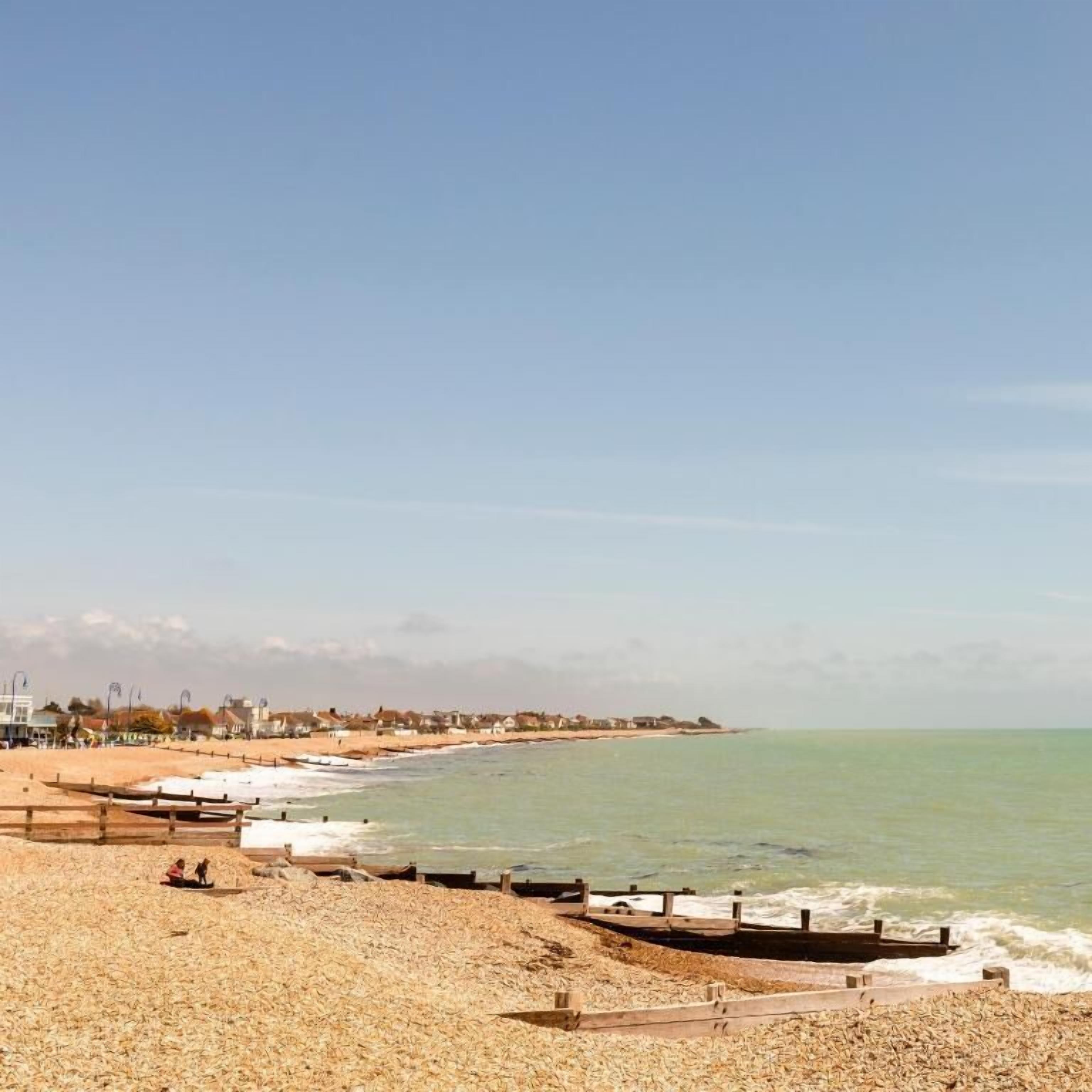 Una playa con un paseo marítimo de madera, vista al océano y casas al fondo.