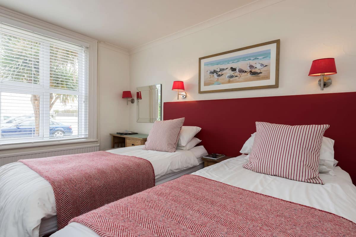A hotel room with two beds, a window with blinds, a painting of birds on the wall, and two wall lamps.