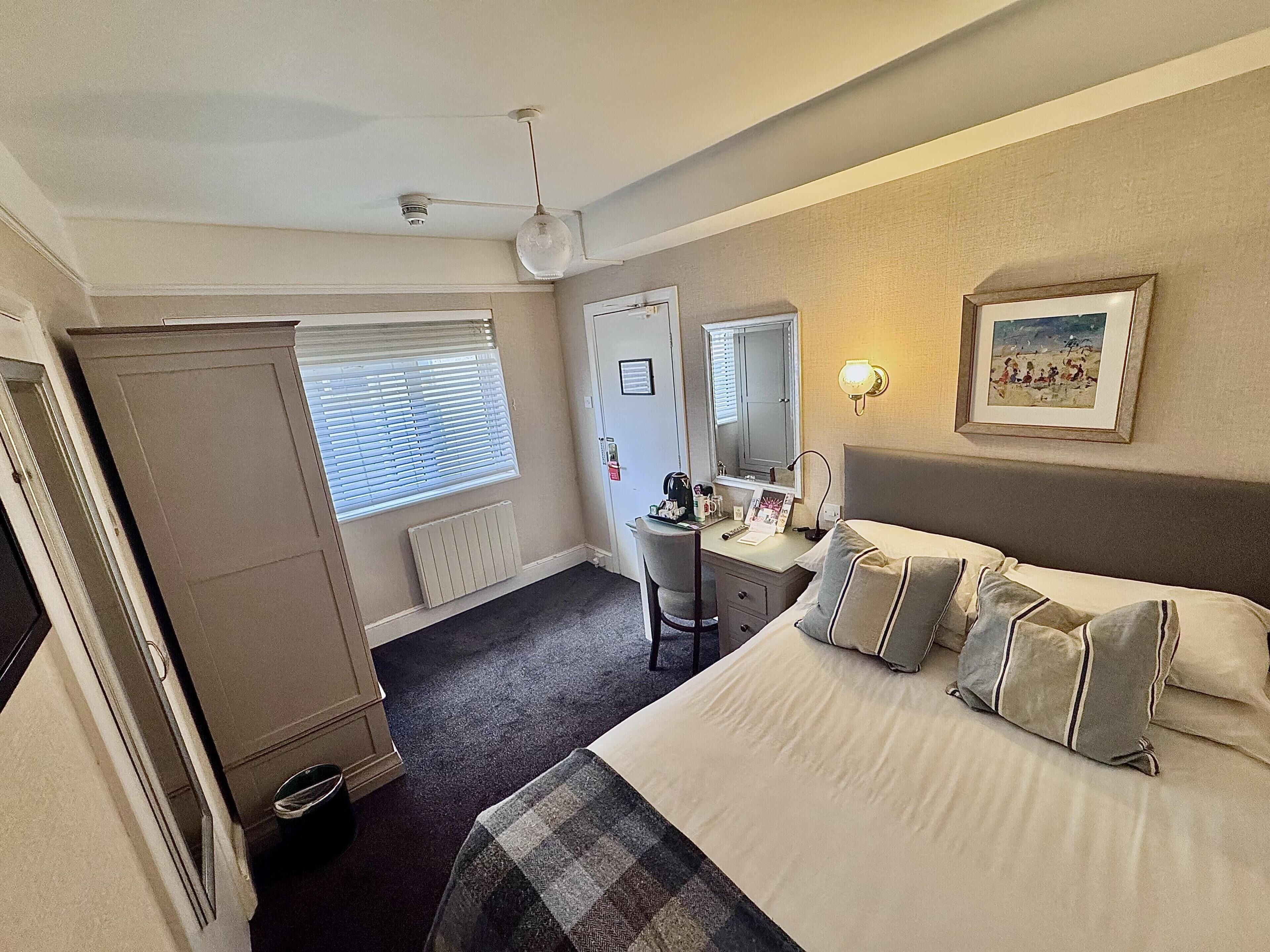 A hotel room with a bed, a desk, a chair, a radiator, a window with blinds, a ceiling fan, and a framed picture on the wall.